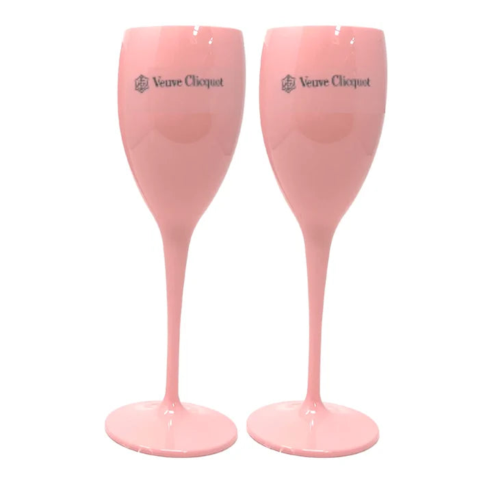 Two individuals holding clear plastic champagne glasses, wearing pink outfits, with one holding a phone and both smiling.