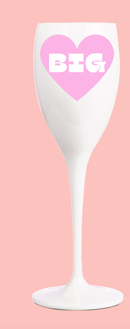 Two individuals holding clear plastic champagne glasses, wearing pink outfits, with one holding a phone and both smiling.