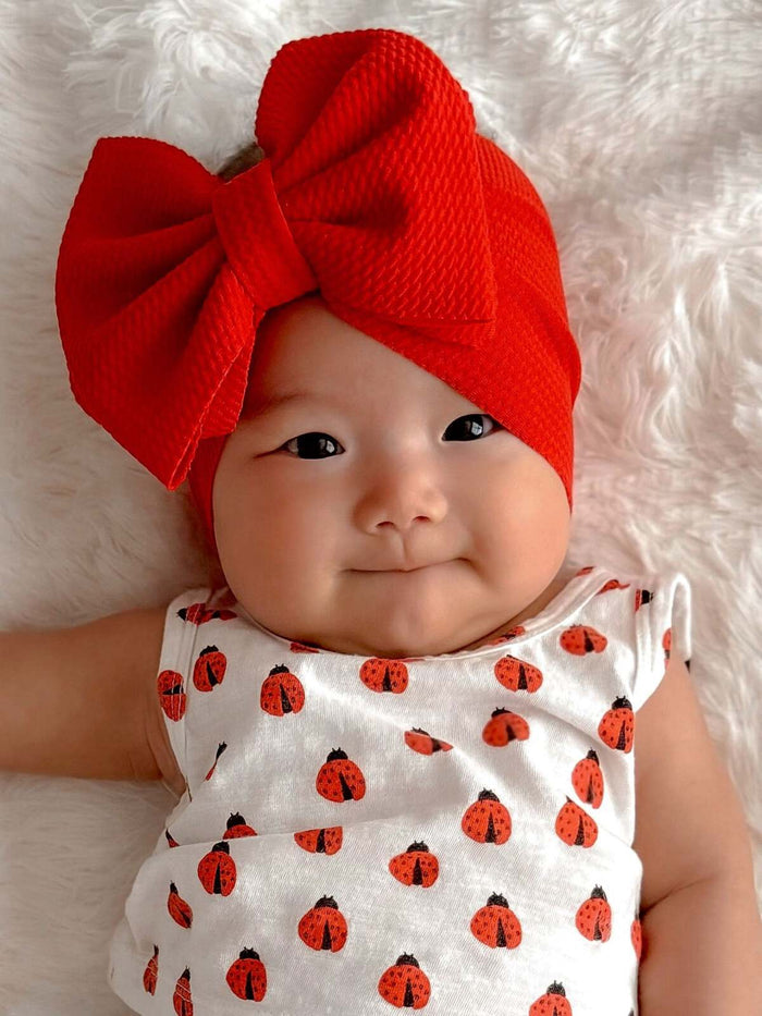 Smiling baby girl in a white ladybug outfit and a large red bow on a soft, textured background.