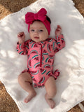 Baby with a pink watermelon-printed swimsuit and a matching headband, lying on a soft white blanket outdoors.