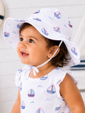 Smiling toddler in a boat-patterned dress and sun hat, with a cheerful expression against a light background.
