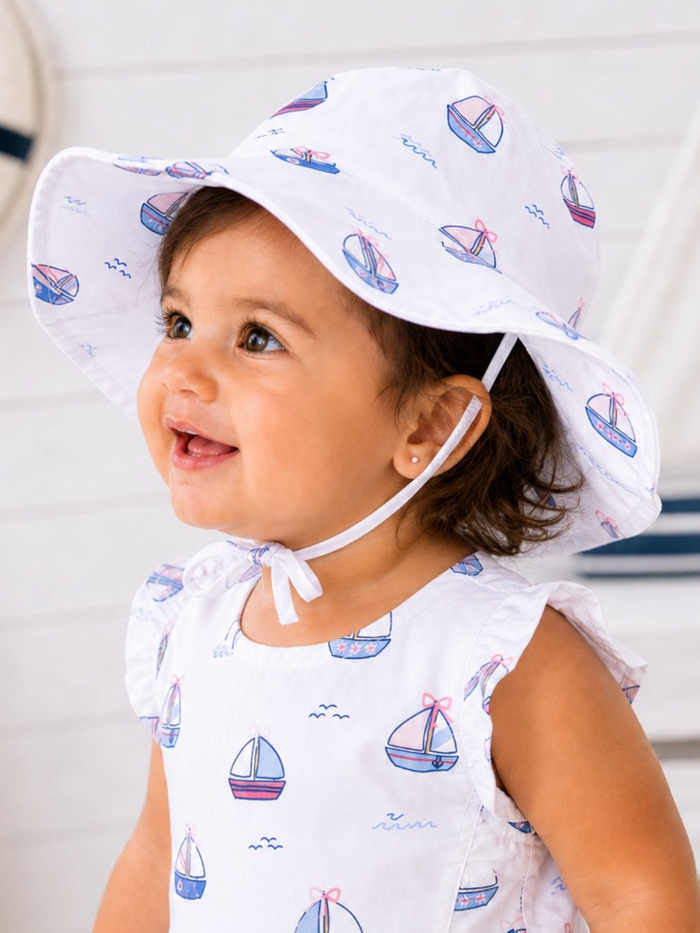 Smiling toddler in a boat-patterned dress and sun hat, with a cheerful expression against a light background.