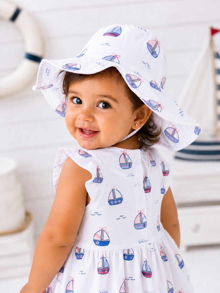 Smiling toddler in a boat-patterned dress and sunhat, with a nautical background.