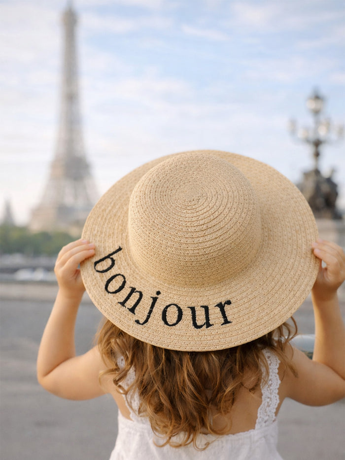 Child wearing a straw hat with "bonjour" text, facing the Eiffel Tower in a scenic outdoor setting.