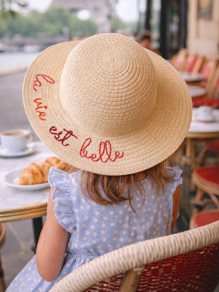 Girl wearing a wide-brimmed straw hat with "la vie est belle" embroidered, sitting at a café table.
