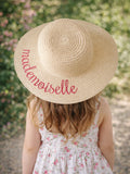 Child wearing a straw sun hat with "mademoiselle" embroidered, surrounded by greenery and floral patterns.
