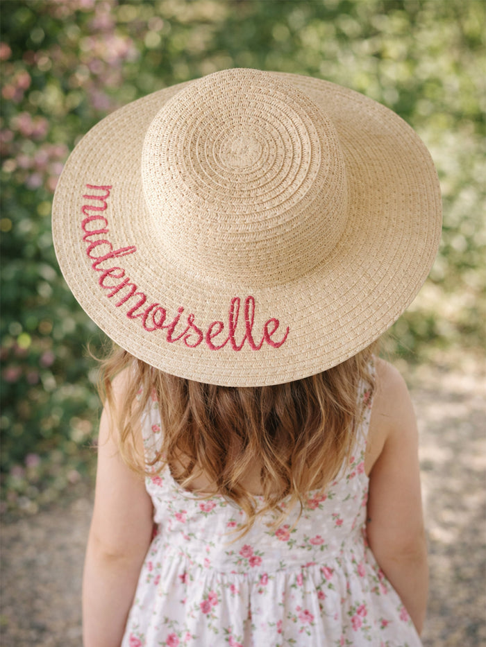 Child wearing a straw sun hat with "mademoiselle" embroidered, surrounded by greenery and floral patterns.