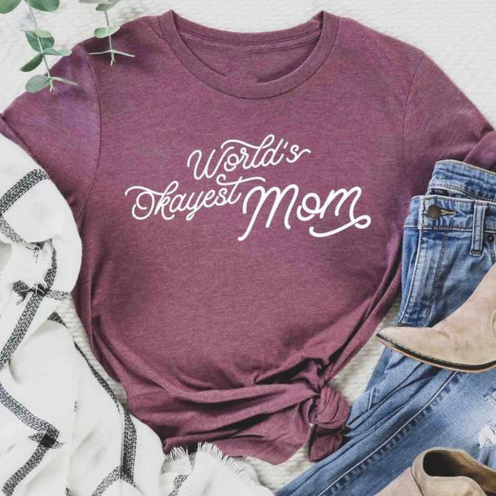 Maroon shirt with "World's Okayest Mom" text, paired with jeans and accessories on a soft surface.