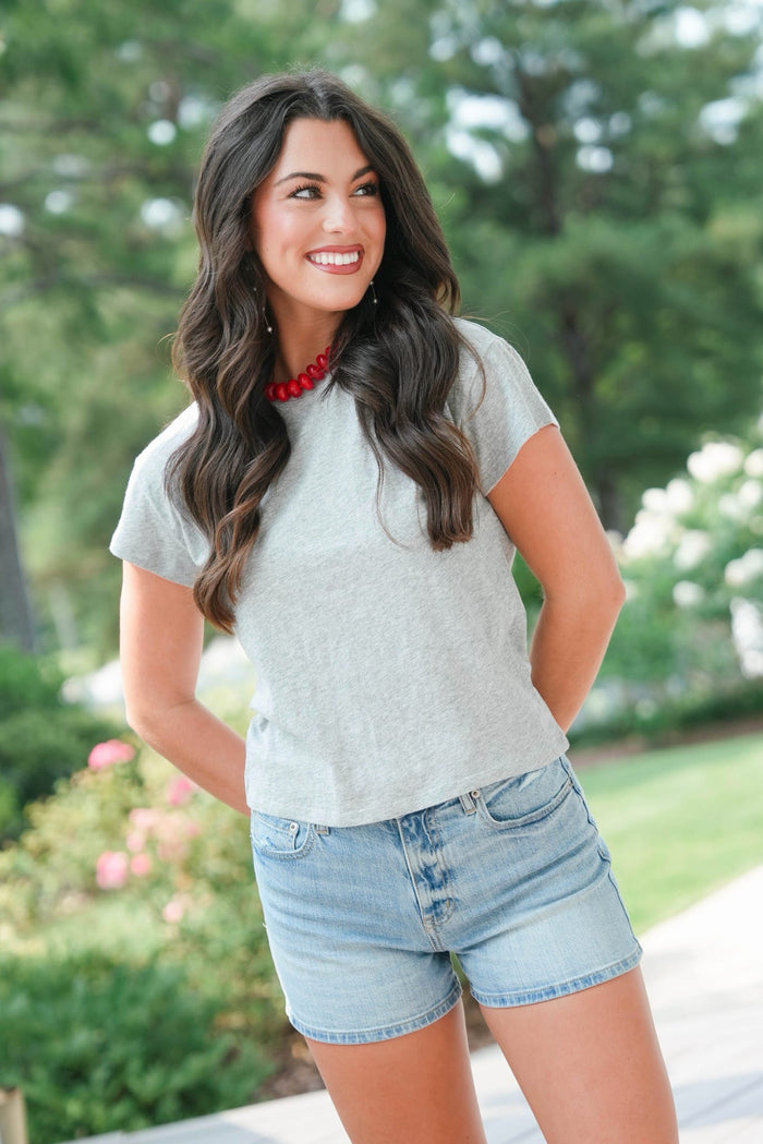Woman wearing a gray t-shirt and denim shorts standing outdoors with greenery in the background